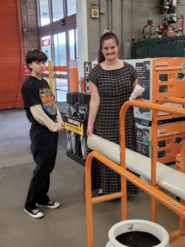 Two students in home depot next to rolling platform, one stabilizing a roll of materials.