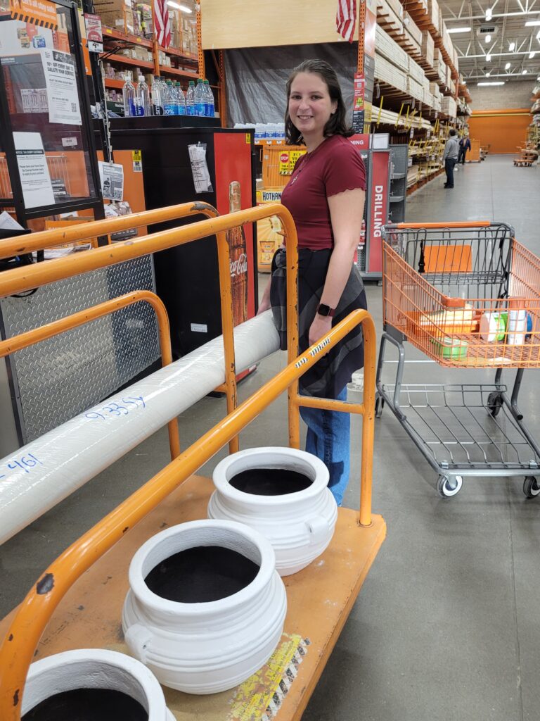 Girl standing in home depot, next to rolling platform with three white pots.