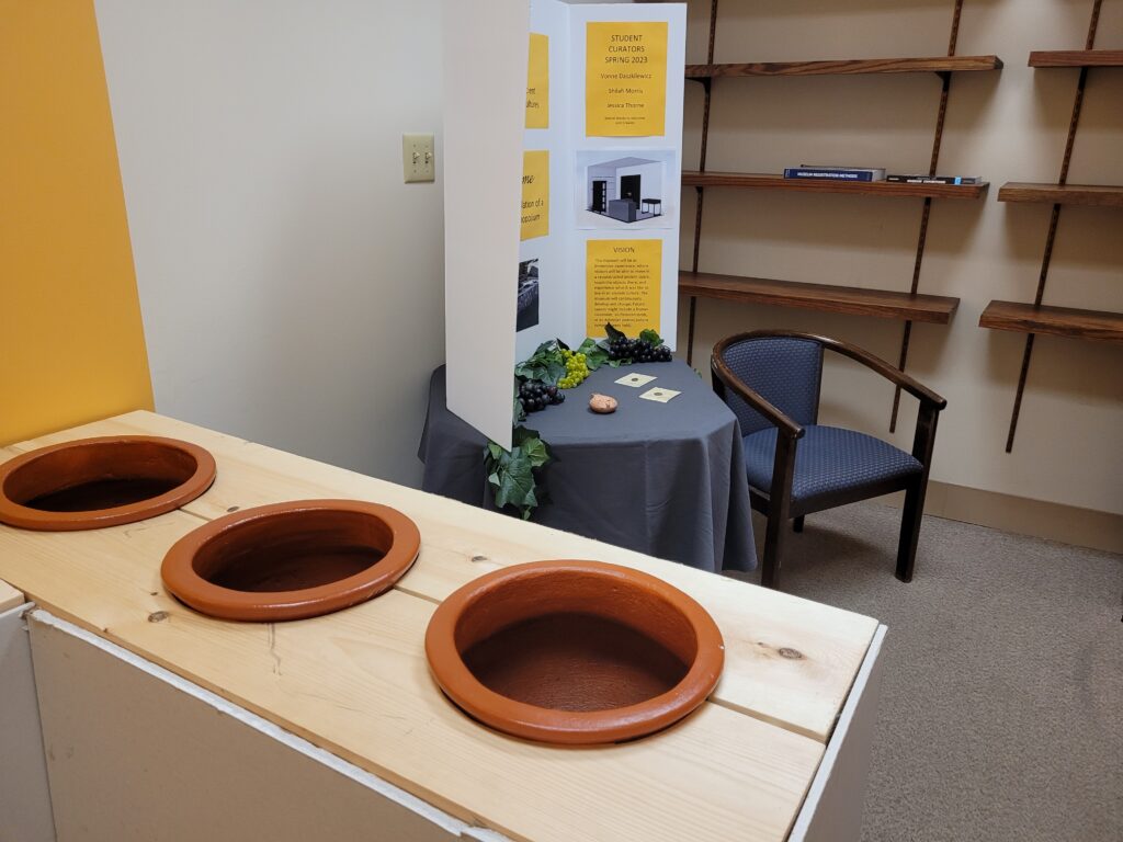 small room consisting of a counter with pots inlayed, chair next to circular table with trifold, and a wall with shelves.