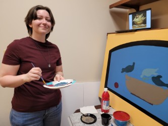 Student holding plate palette next to partially finished painting smiling at camera.